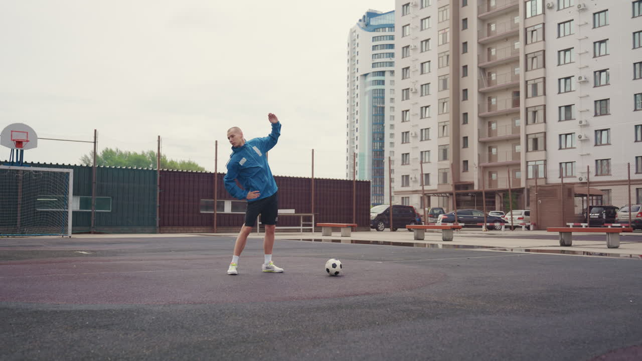 Caucasian player on city court launching juggling drill with apartment highrises backdrop, blue jacket, soccer ball at feet, overcast drizzle, playful confidence, open plaza training, community vibe