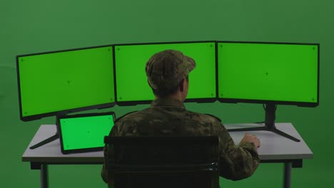 vista trasera de un soldado asiático trabajando con una tableta y varios monitores de computadora con una pantalla verde simulada en un estudio de pantalla verde