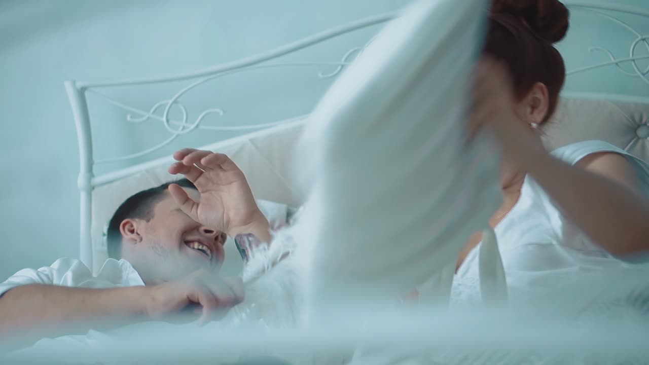 아름다운 젊은 커플이 침대에서 즐겁게 놀고 있습니다. 그들은 베개 에서 싸우고 웃으며 침대에서 카메라를 지나갑니다.