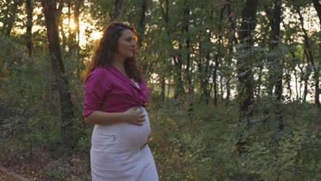 pregnant woman expectant mother walking alone in park forest at sunset, attractive young casual dressed mom stroking caressing touching her belly with hands smiling, medium portrait tracking shot