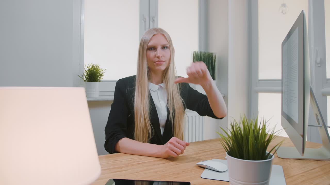 женщина, показывающая большой палец вниз в офисе. сидящая на рабочем месте молодая блондинка в деловом костюме, смотрящая в камеру и делающая большие пальцы вниз жестом неодобрения или неприязни.