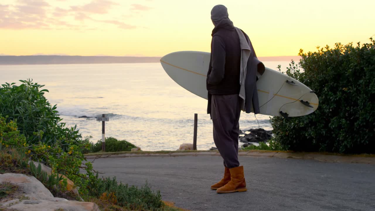 vista trasera de un hombre adulto con una tabla de surf de pie en la carretera cerca de la costa 4k