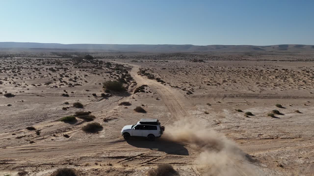 Drone tracking shot of a white off-road vehicle driving through an arid desert landscape on a sunny day, kicking up dust while navigating a curve