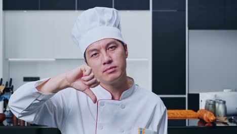 un primer plano de un chef asiático mostrando los pulgares hacia abajo en un gesto a la cámara mientras está de pie en la cocina de su casa