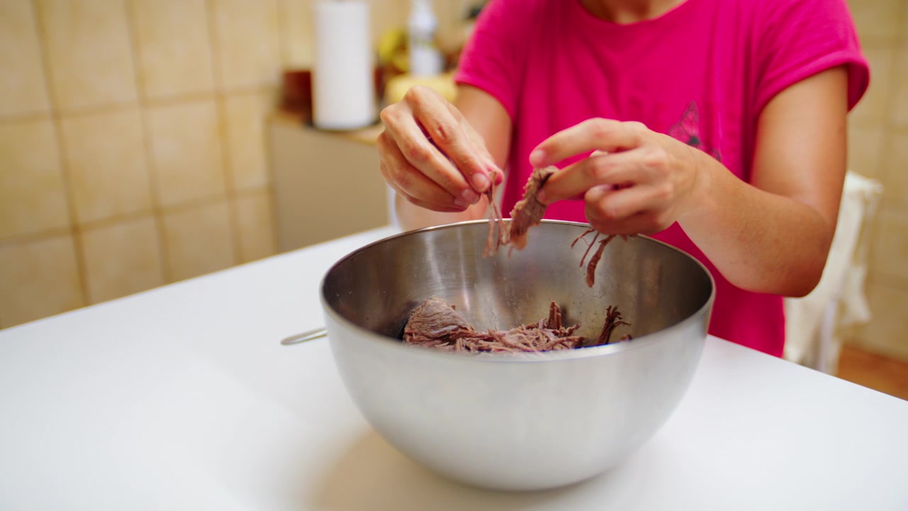 Mani che sfilacciano carne di manzo cotta tenera a mano in una grande ciotola di metallo, preparazione di una ricetta casalinga di manzo sfilacciato