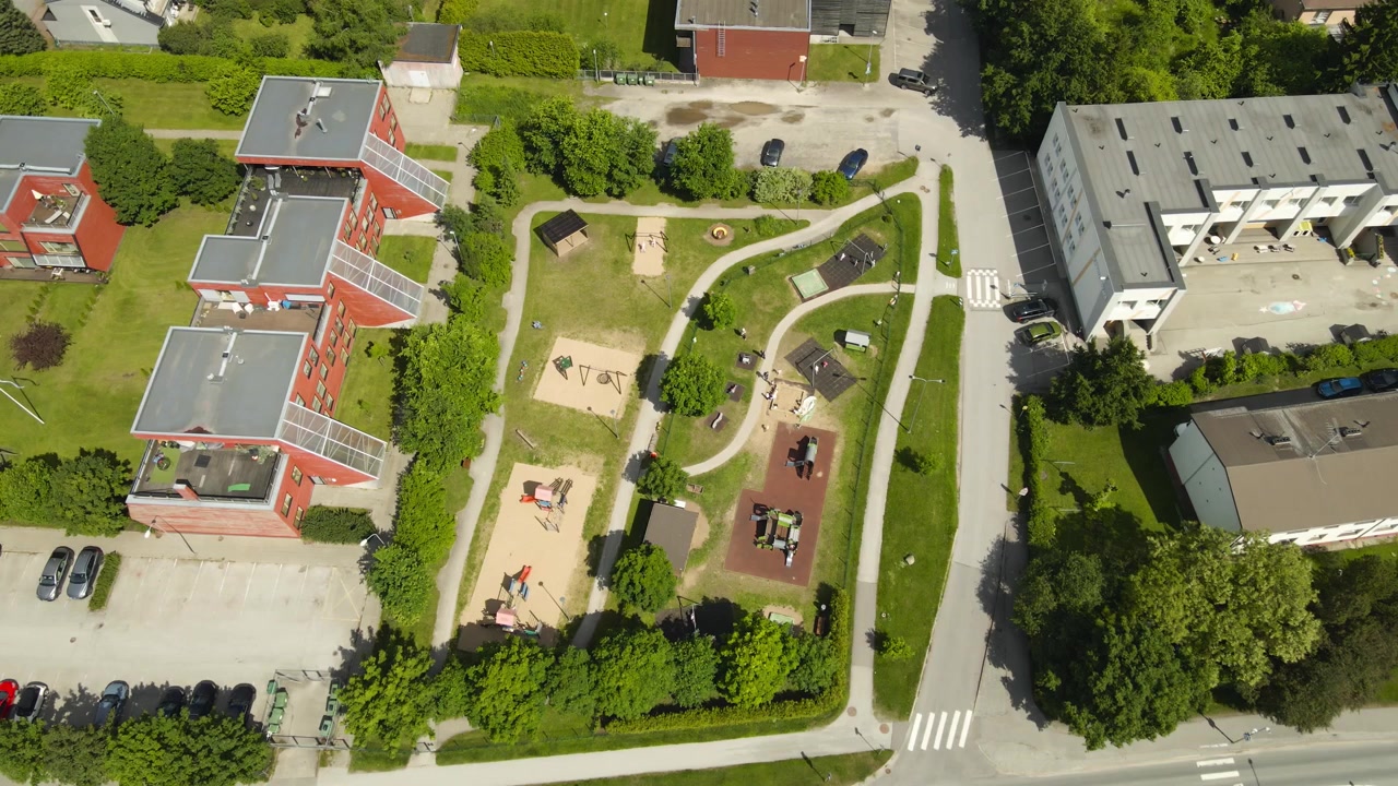 Top down aerial view of a kindergarten playground where children enjoy a sunny summer day, surrounded by residential buildings and lush green trees in a quiet suburban neighborhood