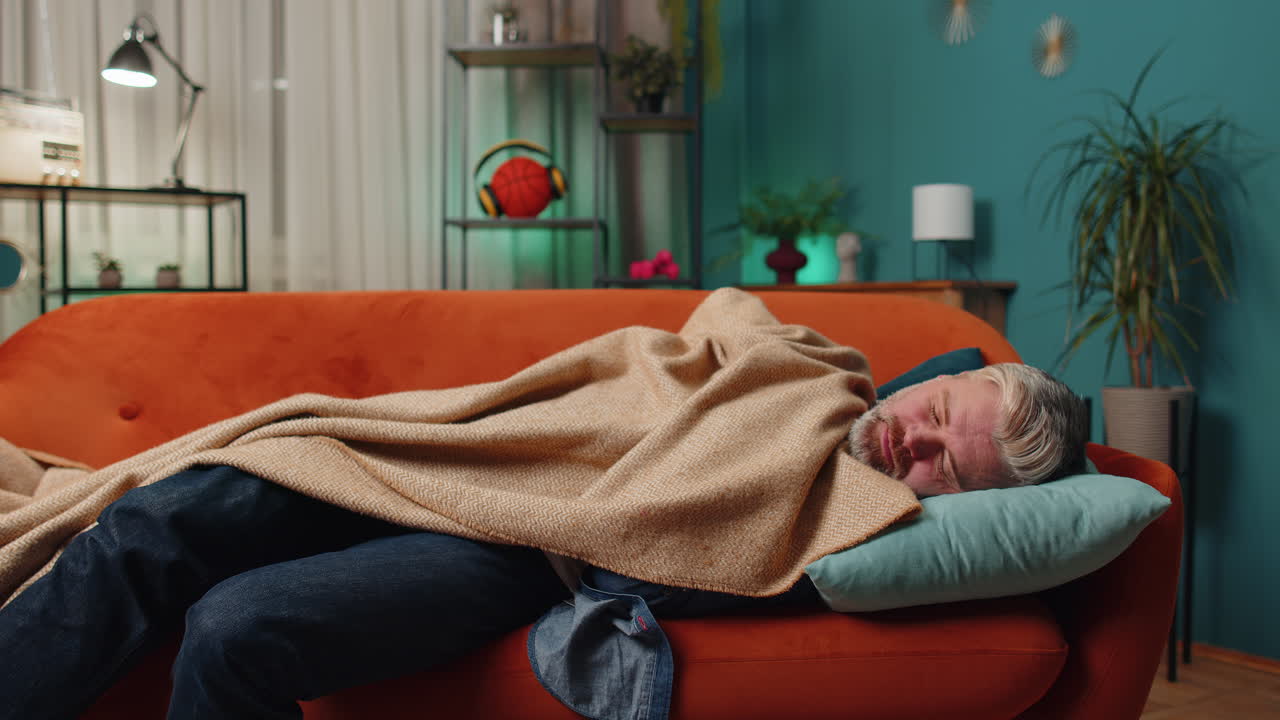 피곤한 중년 성숙한 남자가 침대에 누워 집에서 휴식을 취하고 소파에서 잠들고 있습니다.