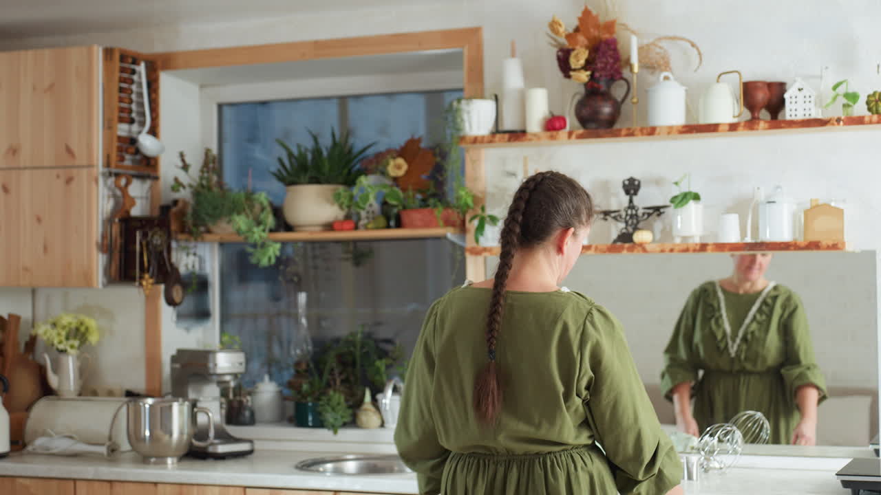 Back view of lady in green dress with long braid walking into cozy kitchen with wooden cabinets, potted plants, kitchenware on shelves, and reflection visible in wall mirror
