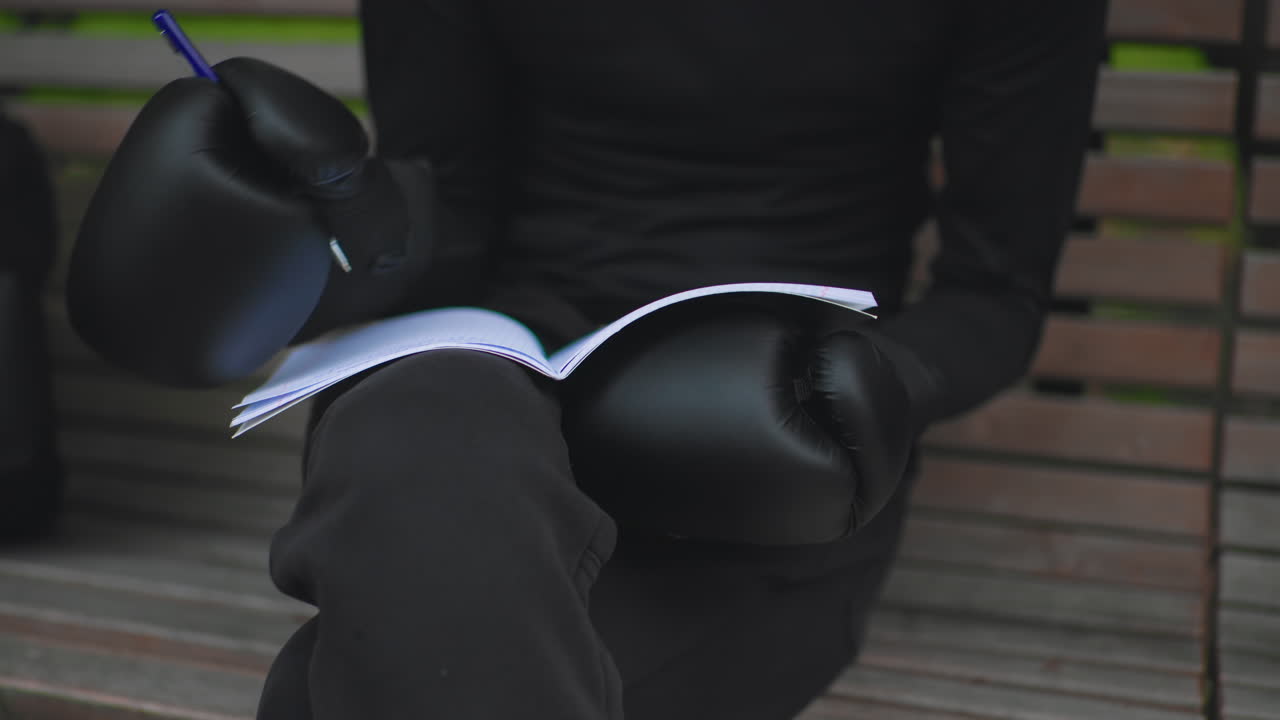 Atleta caucasico con guanti seduto su una panchina del parco che scrive su un foglio, primo piano di muffole di pelle sulle gambe e penna che traccia linee strategiche, atmosfera tranquilla alla luce del giorno, preparazione rituale prima dell'allenamento
