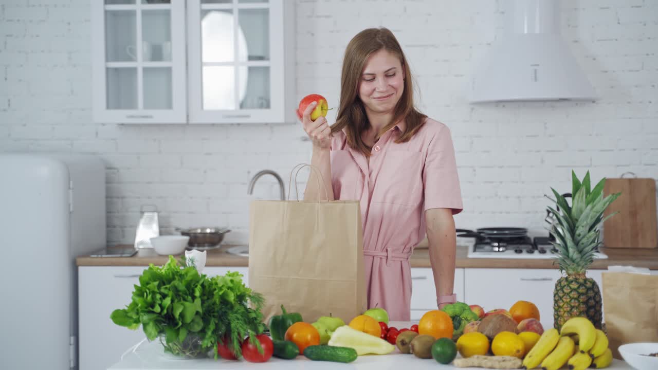 Lovely woman with fresh apple at home. Organic fruit and vegetables on the kitchen table and young female holding an apple. Healthy food.