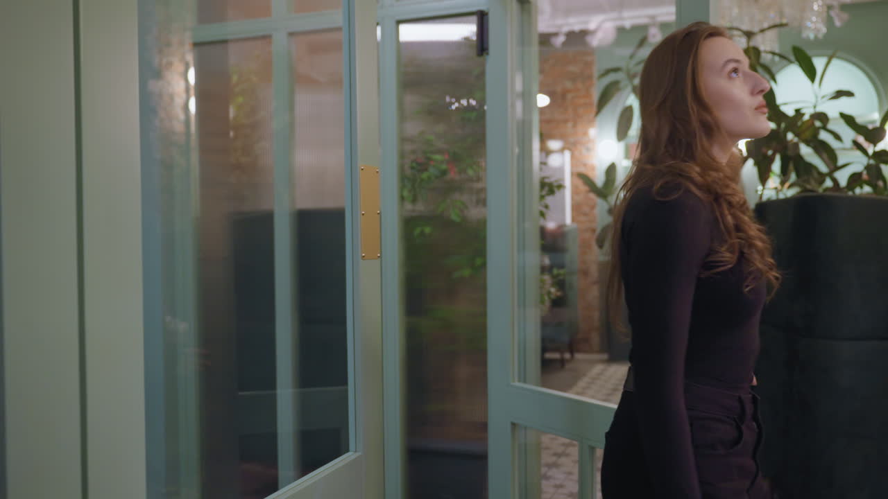 Woman enters cozy lounge through transparent door and gently closes it behind her, surrounded by green cushioned seating and indoor plants in a softly lit, elegant interior