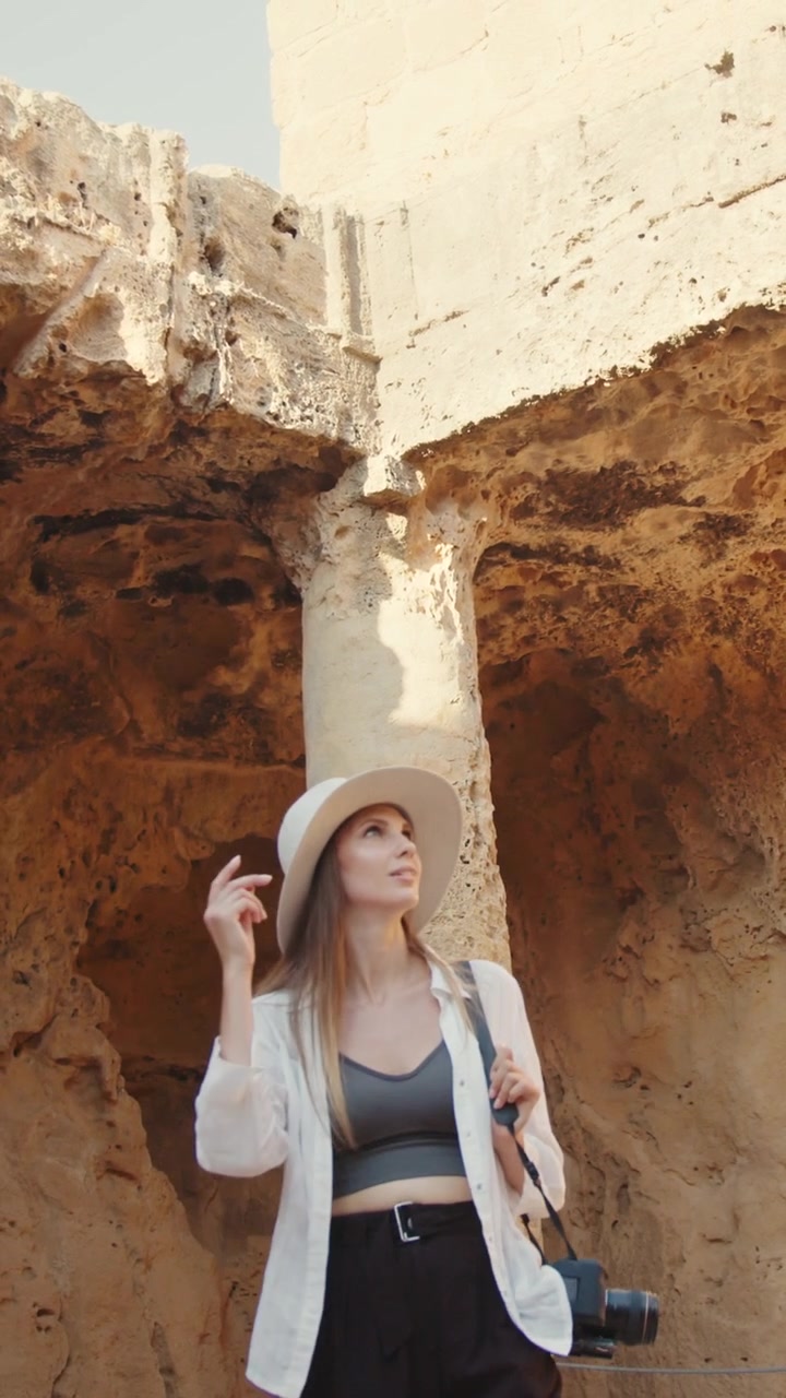 Una donna con un cappello guarda l'architettura di un'antica rovina, tenendo una macchina fotografica e catturando la scena.