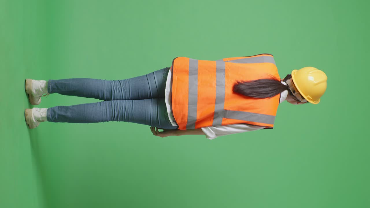 안전 헬을 입은 아시아 여성 엔지니어가 녹색 스크린 배경 스튜디오에 서서 스마트폰으로 이야기하는 전체 몸의 뒷면