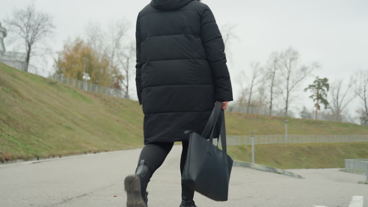 Walking up grassy hill alone. Individual strolling along path with skyline in background. Solitary figure wearing beanie carrying tote walking onto grassy ascent with distant cityscape