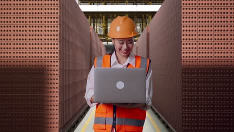 asian female engineer with safety helmet working on a laptop while standing with red brick packed in stacks are stored