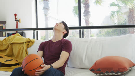 asian male teenager watching tv with basketball and sitting in living room