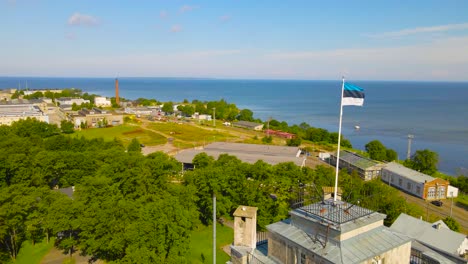aerial orbiting around estonian flag waving in the wind atop the maritime academy building in kopli, tallinn with green treetops on sunny summer day. peninsula view revealing bekkeri port and blue sea