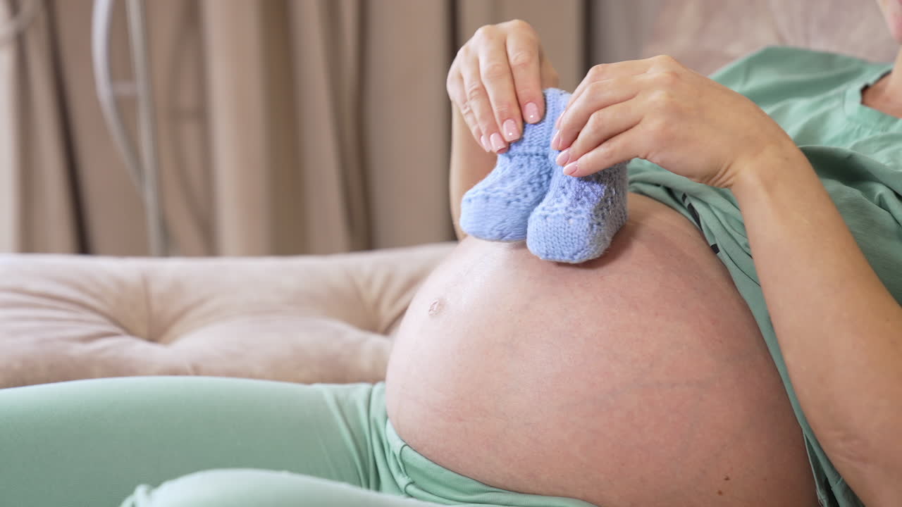 Expectant mother is sitting on the sofa and playing with small baby socks on her big stomach. Woman puts tiny baby booties on the armrest and sets her both hands on the belly to caress it.