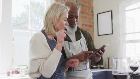 Älteres-Paar-Mit-Gemischter-Abstammung-Trägt-Schürzen-Und-Benutzt-Ein-Digitales-Tablet-Beim-Kochen-In-Der-Küche-Zu-Hause