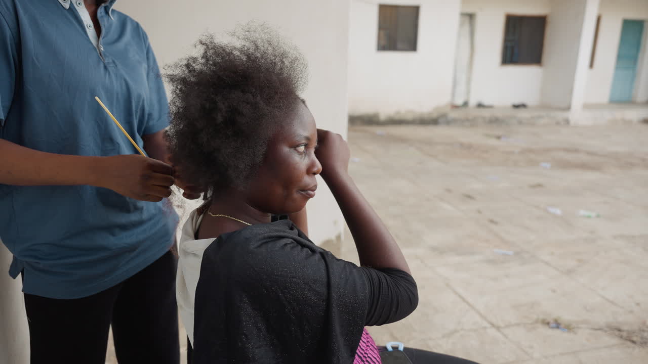 Elderly black woman having hair combed outside, young assistant in blue shirt, gentle hands styling gray hair, courtyard background, relaxed posture, intimate caregiving scene, candid documentary