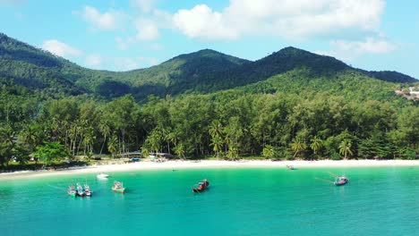 Fishing-boats-anchoring-on-calm-clear-water-of-shallow-turquoise-lagoon-near-white-sandy-beach-of-tropical-island-with-palms-and-lush-vegetation