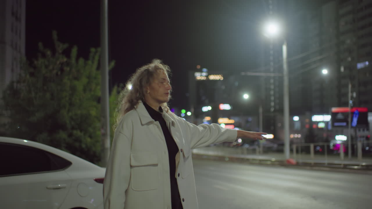 Professional woman stretches out arm to stop car late at night in urban environment with white car parked behind, dressed in oversized coat, illuminated by city streetlights and building signage