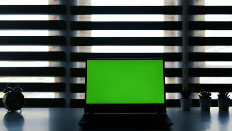 laptop with green screen on white table in front of roller blind. dark office