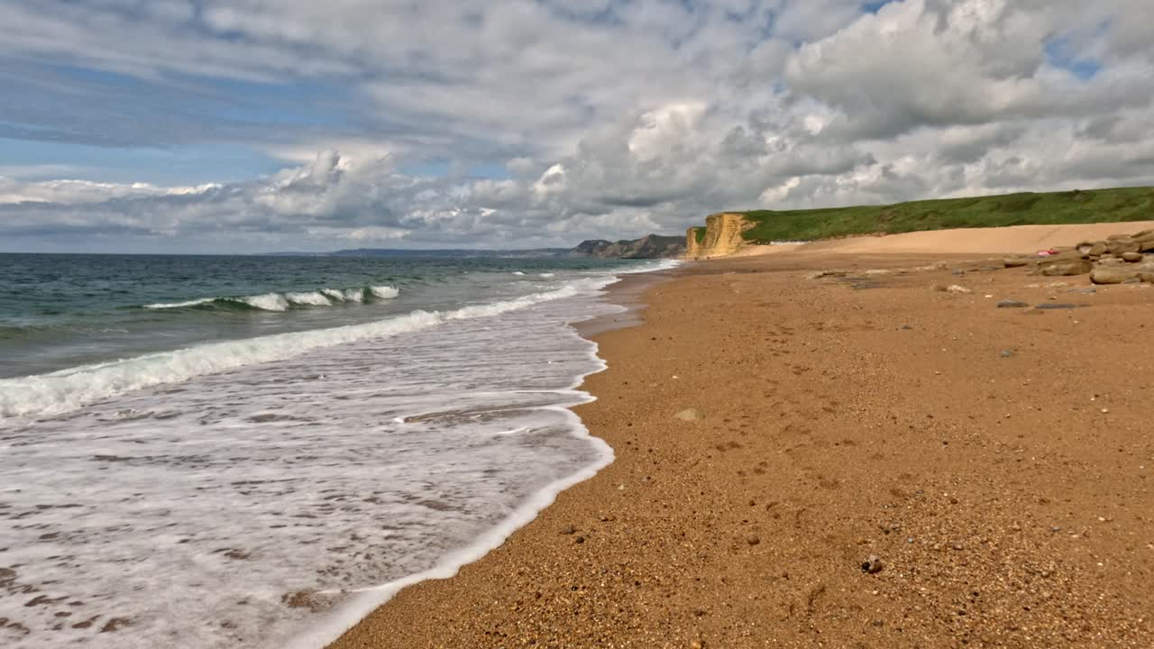 Slow Motion footage of the waves crashing up onto a beach on the Jurassic Coast, Dorset. The footage is low and moves backwards with amazing cliffs in the background and a sky full of clouds above.