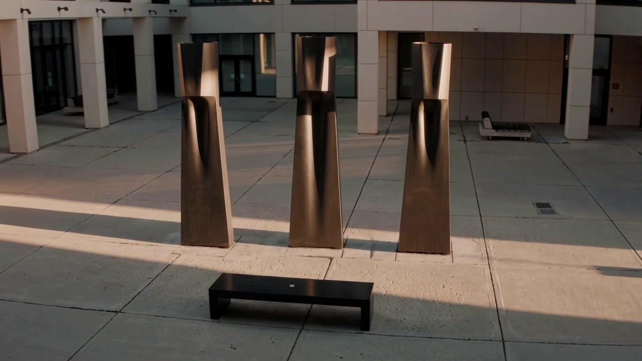 La telecamera arretra rivelando un trio di sculture in bronzo e una panchina in piazza, mostrando i dettagli dell'edificio