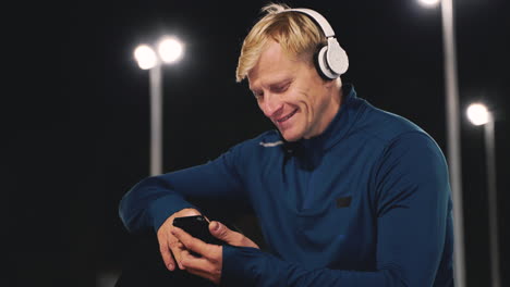 deportista feliz sentado en el parque escuchando música con auriculares bluetooth y usando el teléfono móvil mientras toma un descanso durante su sesión de entrenamiento por la noche