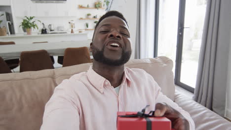 african american man with red gift box smiles, blows a kiss in a bright living room