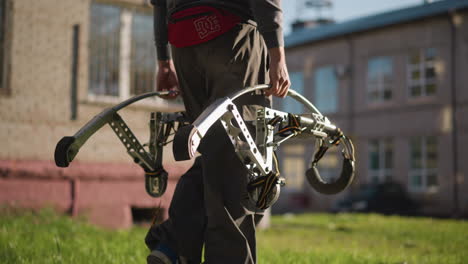 a guy carrying large metallic spring stilts, wearing black pants, dark gray long-sleeved shirt, and red hip bag with visible white logo. background is blurred with grassy area and distant building