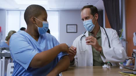 Caucasian-male-doctor-giving-covid-vaccination-to-african-american-female-doctor,-both-in-face-masks