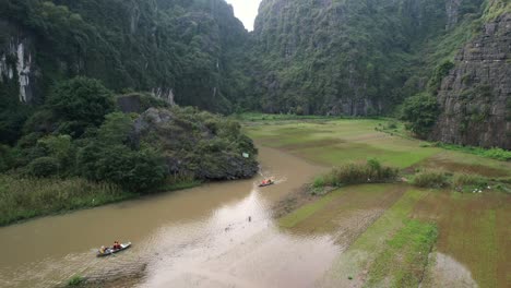 Barcos-Turísticos-Sampán-Con-Turistas-Navegan-Por-El-Río-Ngo-Dong-Corriendo-En-Un-Barranco-De-Piedra-Caliza-En-El-Parque-Nacional-Ninh-Binh,-Campos-De-Arroz-Inundados-En-Las-Orillas-Del-Río---Ascendente-Aéreo