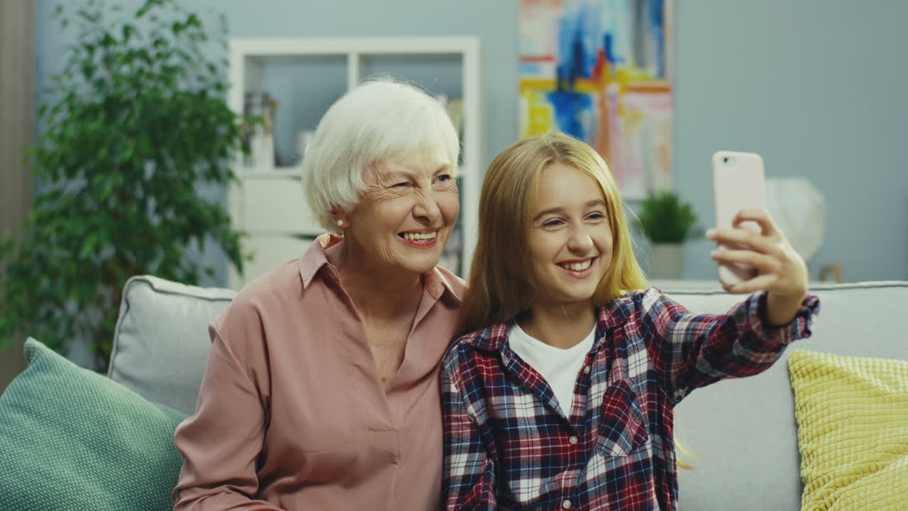 아늑한 거실에서 할머니와 함께 소파에 앉아 스마트폰 카메라로 셀카 사진을 찍는 귀여운 십대 소녀의 초상화