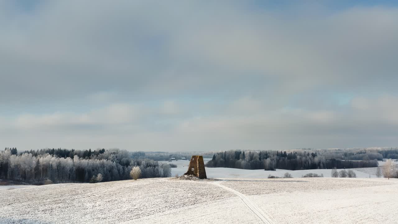Aerial drone shot of ancient windmill ruins on a small hilltop in the countryside. Snow covered ground and frozen forest in the background on a sunny winter day.