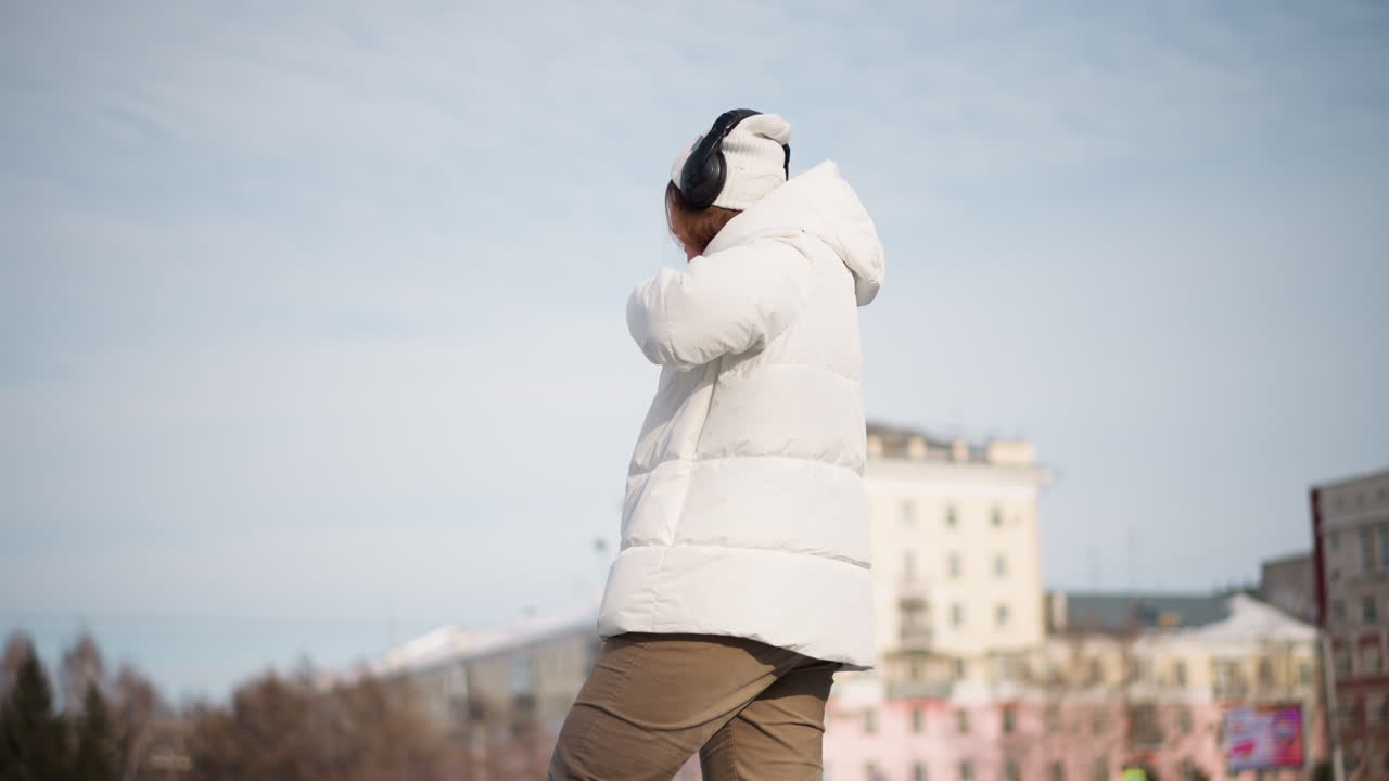 Back view girl wearing white puffer coat, beanie and headphones practices kung fu dance moves on tiled snowy plaza in urban park under clear winter daylight, expressing fluid martial art style