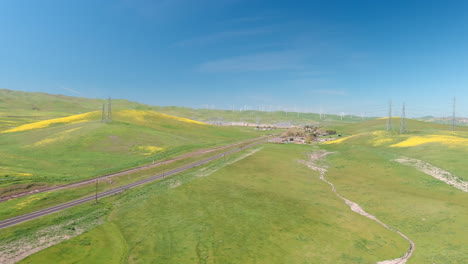 large powerlines cross a highway as wind turbines spin in the background on green hills dotted with yellow wildflowers, aerial