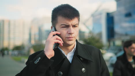 close-up of man in black coat answering phone call while outdoors in urban setting, background features blurred modern buildings, greenery, and passerby, man appears focused, dressed professionally