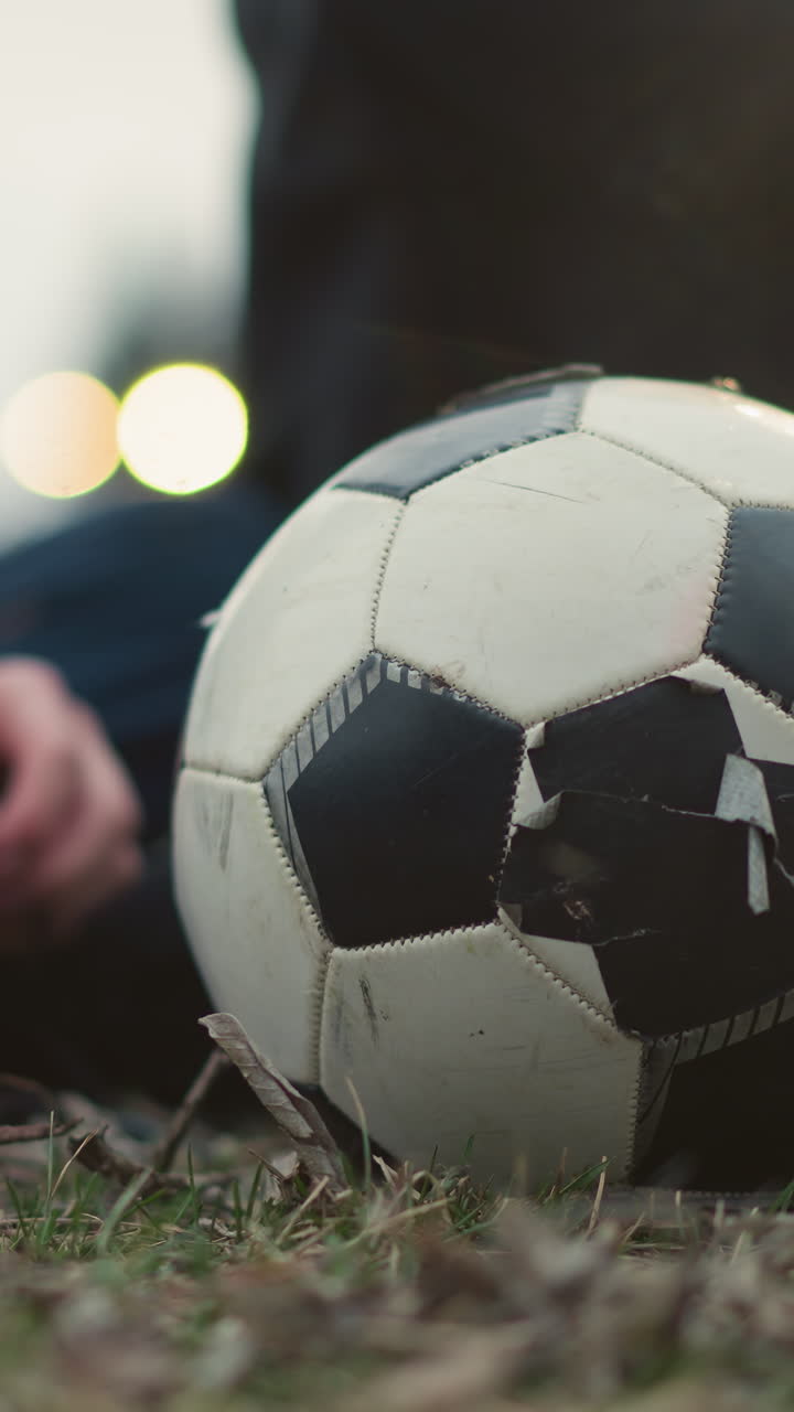 Close-up of a worn soccer ball on a grassy field with two people seated behind it, one of them in a black outfit appears to be picking something from the ground