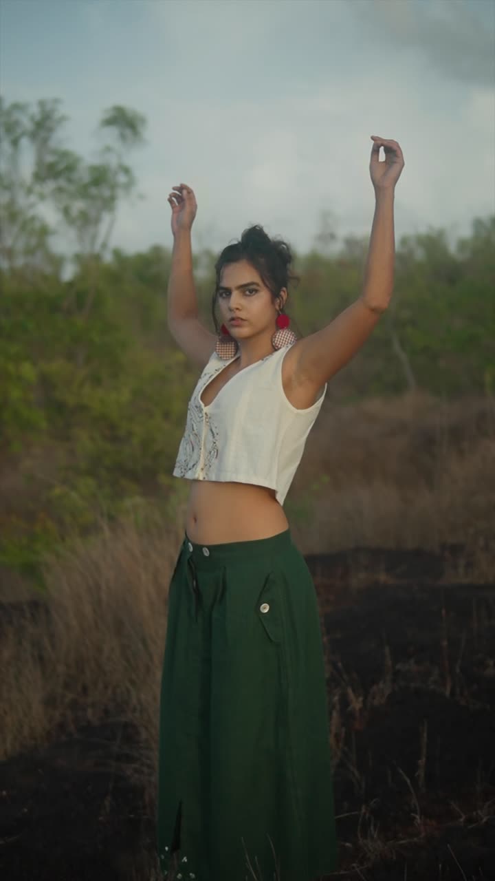 Woman in a white cropped top and green palazzo pants walks through a dry, open landscape with a confident gaze