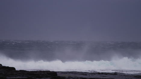 strong offshore winds lift the lip of a wave crashing onto the rocks in slow motion during a storm