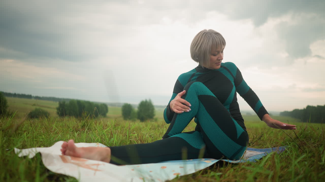 요가 매트 위에 앉아있는 중년 여성은 차분한 표정으로 앉아서 회전하는 연습을 하고 있으며, 천천히 머리를 뒤로 들어 올리고, 거대한 풀과 구름이 가득한 하늘 아래 멀리 있는 나무로 둘러싸여 있습니다.