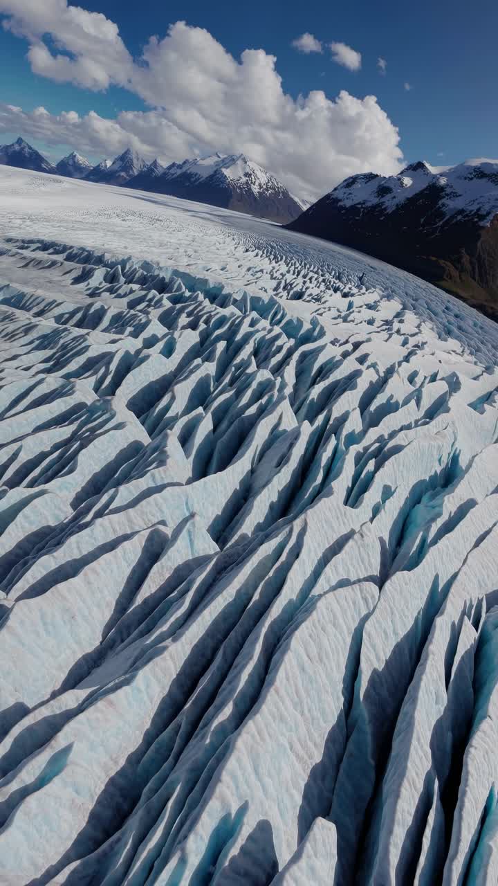 Aerial view of a vast glacier with dramatic crevices, set against a backdrop of snowy mountains