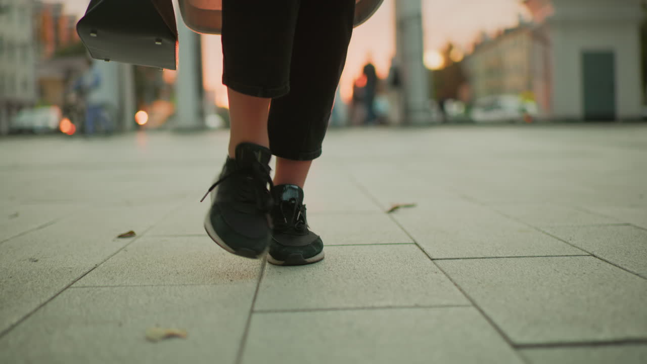 leg view of person walking on tiled pavement wearing black canvas sneakers and carrying black handbag during sunset with dry leaves scattered on ground