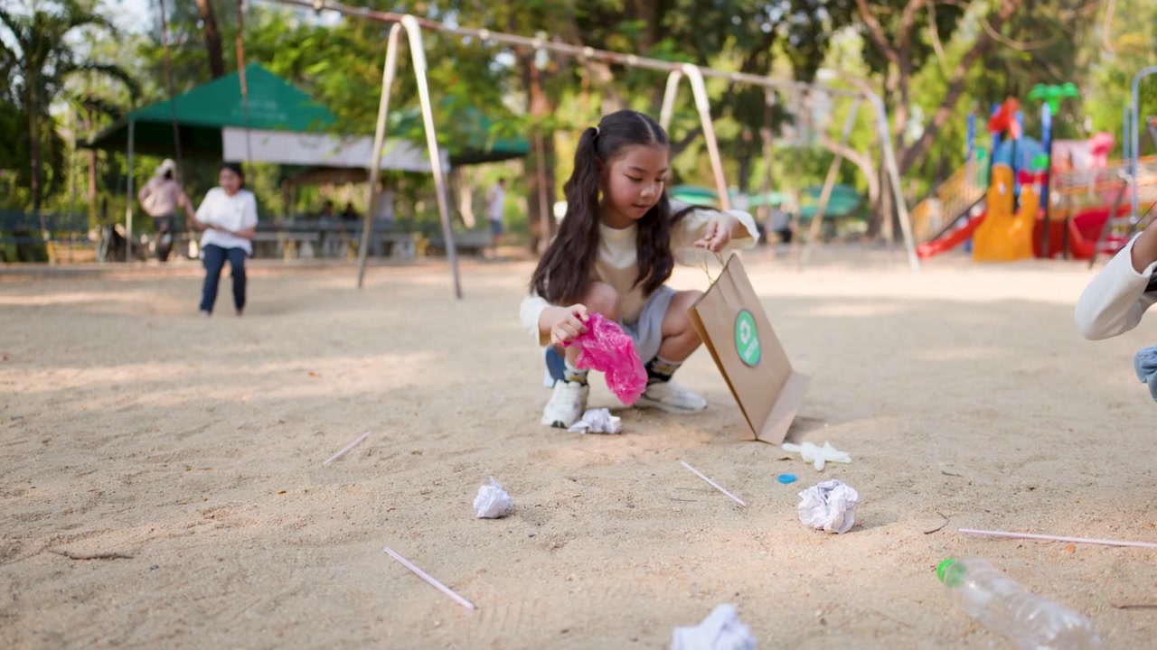 Two Asian girls collect litter into paper recycle bags at a sandy park playground.