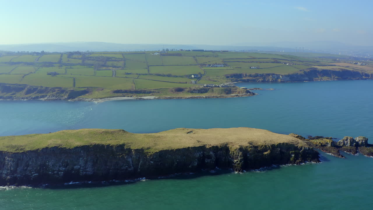 Dramatic aerial perspective of Portmuck Island's rugged shoreline meeting azure waters, with Antrim's distant coastline framing the horizon.