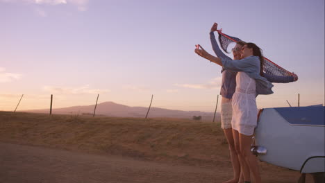 beautiful girl friends taking selfies on road trip at sunset with vintage car