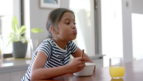 Biracial-girl-eating-breakfast-cereal-sitting-at-island-in-sunny-kitchen,-copy-space,-slow-motion