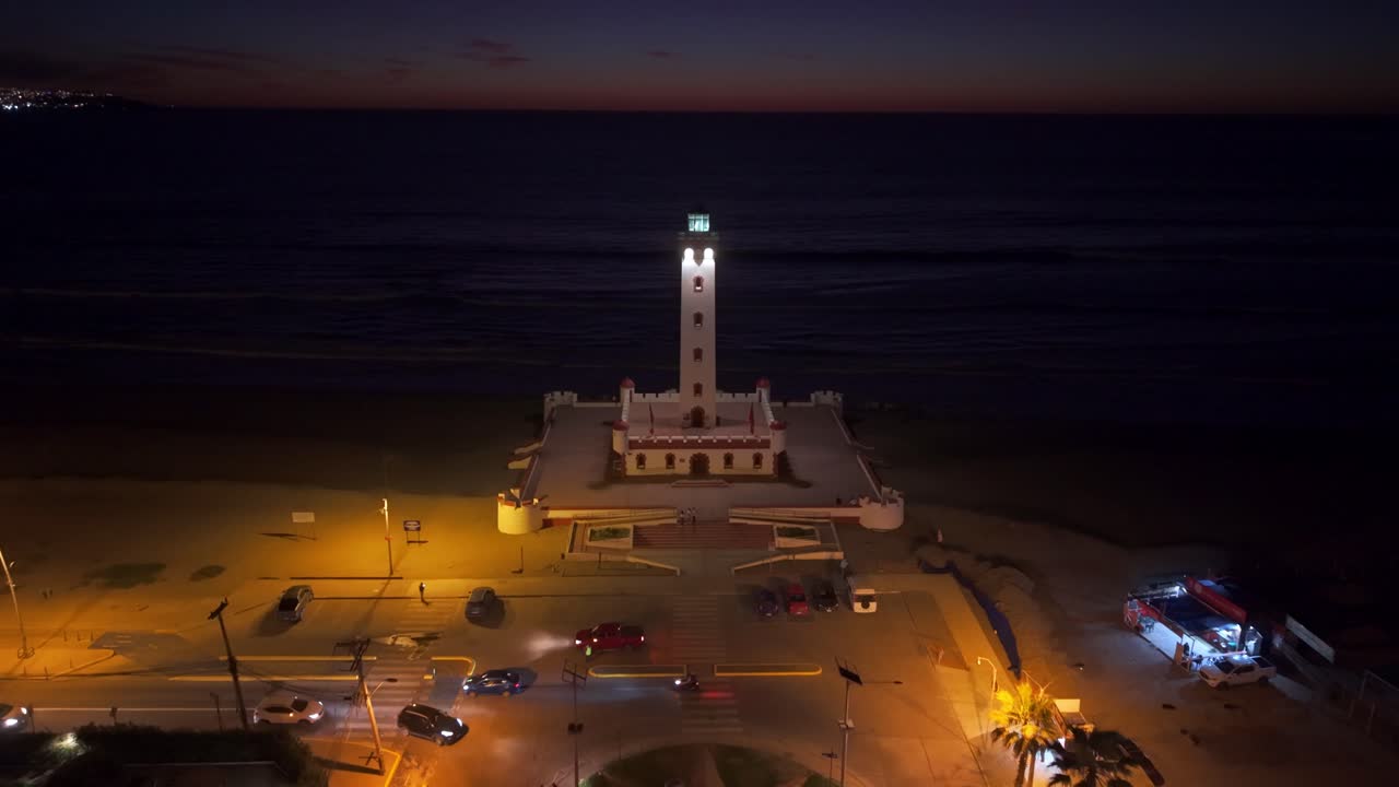 Aerial view establishing tilt up of the monumental lighthouse of La Serena at night with lights and the silhouette of the sunset in its final phase in the background